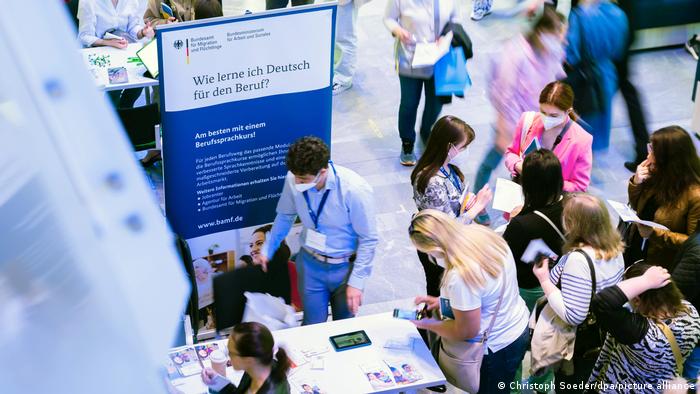 Foto von einer Jobmesse in Berlin. Teilnehmer stehen an Infoständen. Auf einem Aufsteller ist zu lesen «Wie lerne ich Deutsch für den Beruf?». 