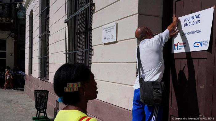 Voluntad de elegir. 27 de noviebre de 2022, pone en el cartel que un operario pega en la puerta de un edifico oficial ante la mirada de una transeúnte.