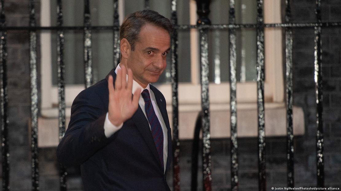 Kyriakos Mitsotakis, in a dark suit, raises his hand in a wave on a visit to London last year.