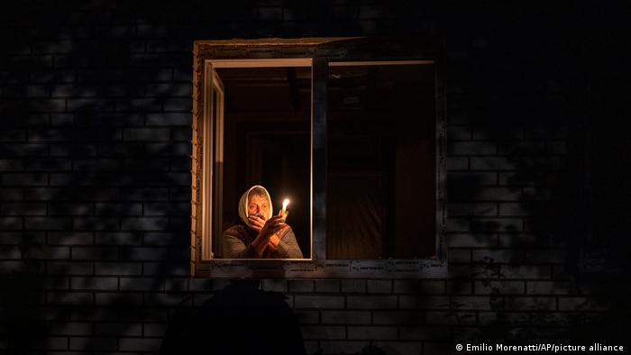 Una mujer sostiene una vela, asomada a la ventana, en medio de la oscuridad.