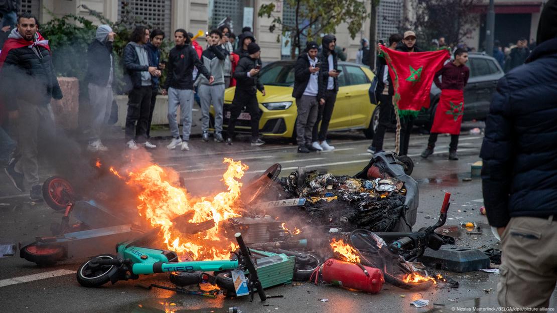 Moroccan men, some draped in and displaying national flags, stand next to a burning heap of e-scooters in Brussels, Belgium