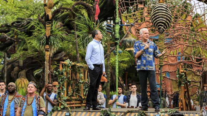 James Cameron steht mit einem Mikro in der Hand vor einer Kulisse, die einem Urwald ähnelt.