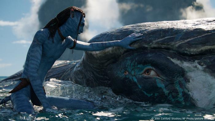 Eine blauhäutige humanoide Person streichelt einen Wal.