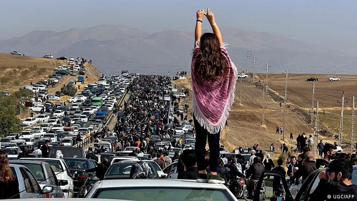 A back view of a woman with loose hair stands on the roof of a car and stretches her arms in the air.  The car is part of a protest convoy of hundreds of other cars in a steppe-like landscape.