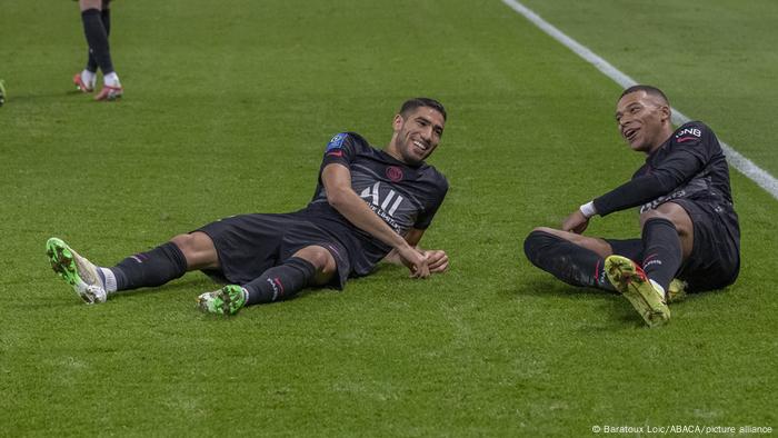 Los jugadores del PSG Achraf Hakimi y Kylian Mbappé se ríen en el césped.