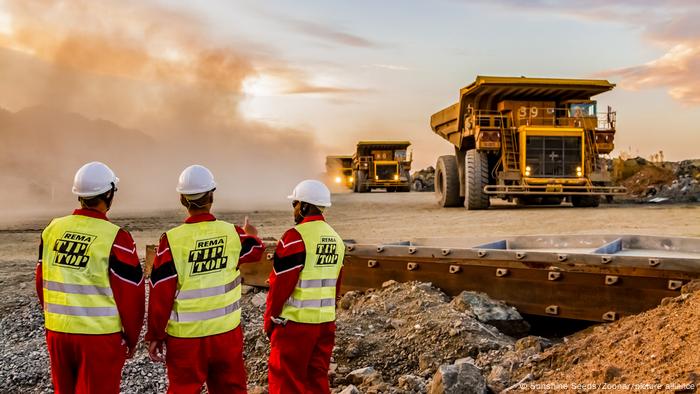 Drei Männer in Bauarbeiterkleidung schauen auf große LKW, die Platin-Erz aus einer Mine in Südafrika abtransportieren