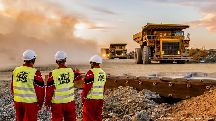 Drei Männer in Bauarbeiterkleidung schauen auf große LKW, die Platin-Erz aus einer Mine in Südafrika abtransportieren Drei Männer in Bauarbeiterkleidung schauen auf große LKW, die Platin-Erz aus einer Mine in Südafrika abtransportieren