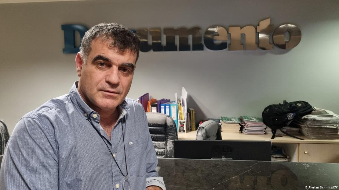 Kostas Vaxevanis, in a grey shirt, sits in front of a desk with the 'Documento' logo above it on the wall