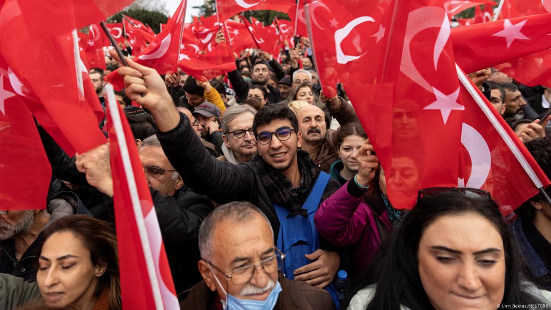 Anhänger des Istanbuler Bürgermeisters Ekrem Imamoglu versammeln sich bei einem Protest in Istanbul. Eine große Menschenmenge, vorne sind Frauen und Männer mit rot-weißen türkischen  Fahnen. In der zweiten Reihe schwenkt ein junger Mann mit Brille auch eine Fahne 