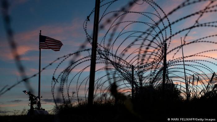Eine US-Flagge weht neben einem Zaun aus mehreren Stacheldraht-Rollen an der US-mexikanischen Grenze.