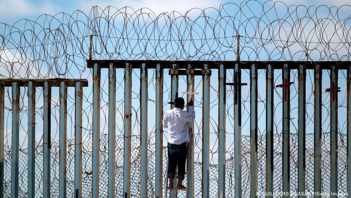 Ein Mann hängt ein Holzkreuz an eine der senkrechten, rostigen Stahlstreben des Stacheldraht-bewehrten Grenzzauns bei Tijuana, im Hintergrund ist der blaue Himmel zu sehen.