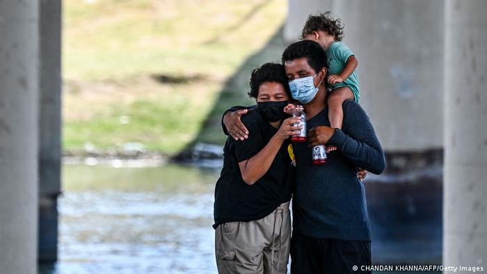 Ein Paar, beide mit Mundschutz, umarmt einander, nachdem es den Rio Grande durchquert hat. Der Mann trägt ein Kleinkind auf seinen Schultern. 