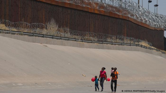 Eine Familie (Vater, Mutter, zwei Kinder) geht auf der Südseite an dem hoch aufragenden Grenzzaun zu der USA entlang