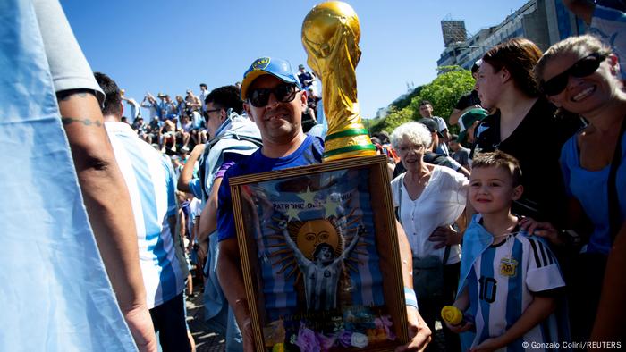 Un hombre posa con una réplica de la Copa del Mundo y un cuadro con la imagen de Diego Armando Maradona.