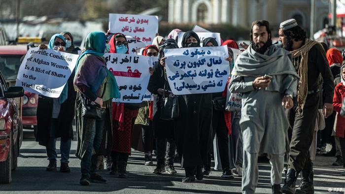 Eine Gruppe Frauen hält Transparente auf einer belebten Kabuler Straße in die Höhe. Rechts im Bild sind zwei Männer zu sehen, die den Protest misstrauisch zu beobachten scheinen.