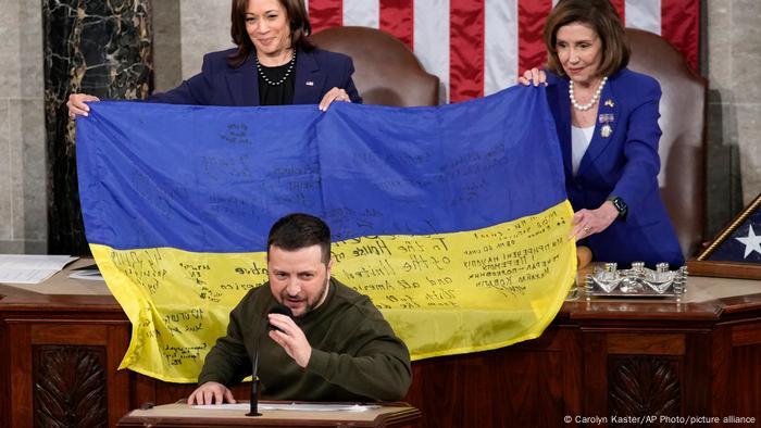 Kamala Harris, vicepresidenta de Estados Unidos, y Nancy Pelosi, presidenta de la Cámara de Representantes, sonríen detrás de un escritorio. Sostienen una bandera ucraniana con una inscripción detrás del presidente ucraniano Zelenski, que habla por un micrófono,