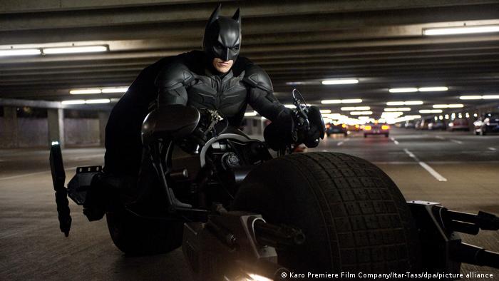Batman sitzt auf einem großen Motorrad mit sehr dicken Reifen und fährt durch eine Tiefgarage.