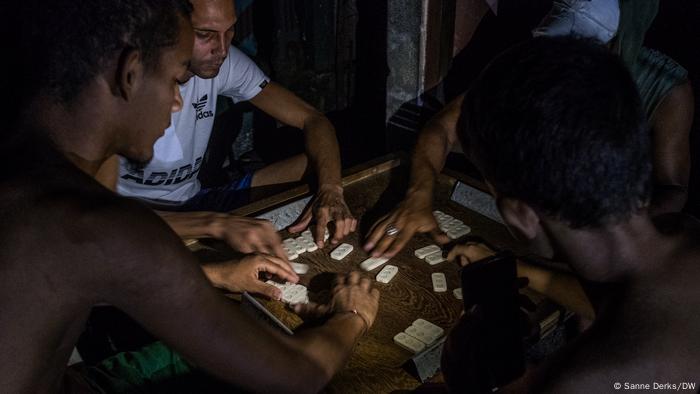 Un grupo de tres personas jugando al dominó, con el tablero iluminado por un teléfono móvil.