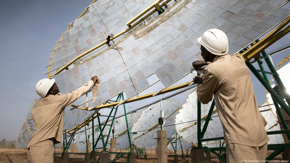 Two workers wearing white helmets and beige suits adjust solar energy installations.