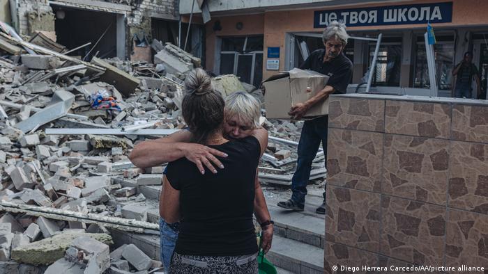 Dos profesoras se abrazan delante de una gran pila de ladrillos en ruinas. Un hombre saca una caja de lo que queda del edificio, que tiene un cartel que dice ESCUELA en cirílico.