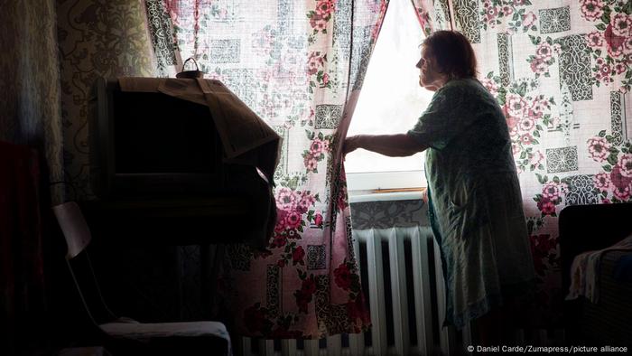 Una anciana en un apartamento poco iluminado aparta unas finas cortinas floreadas para mirar por la ventana.