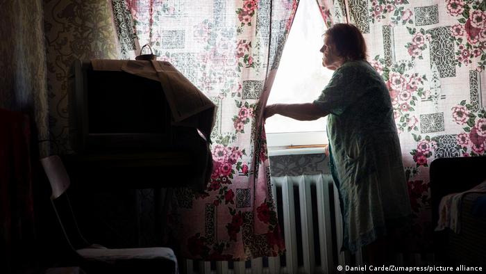Una anciana en un apartamento poco iluminado aparta unas finas cortinas floreadas para mirar por la ventana.