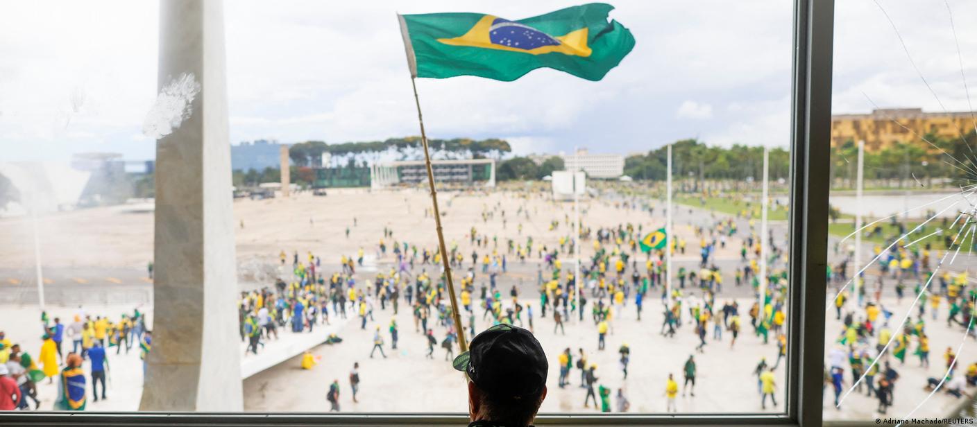Em primeiro plano, homem de costas veste uma camisa amarela que diz "Deus acima de tudo". Ele observa uma multidão de pessoas vestindo verde e amarelo em um espaço aberto à sua frente. Uma bandeira do Brasil está hasteada bem no centro.