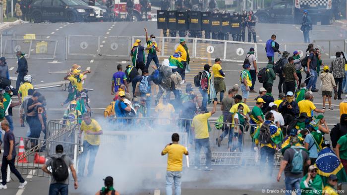 Tumult unter Bolsonaro-Anhänger überwiegend in brasilianischen Farben gekleidet, Rauch ist zu sehen und in einiger Entfernung ein knappes Dutzend Polizisten in Schutzausrüstung