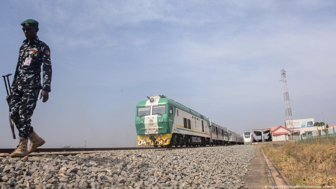 An armed man walking along railroad tracks with a train and station in the background An armed man walking along railroad tracks with a train and station in the background