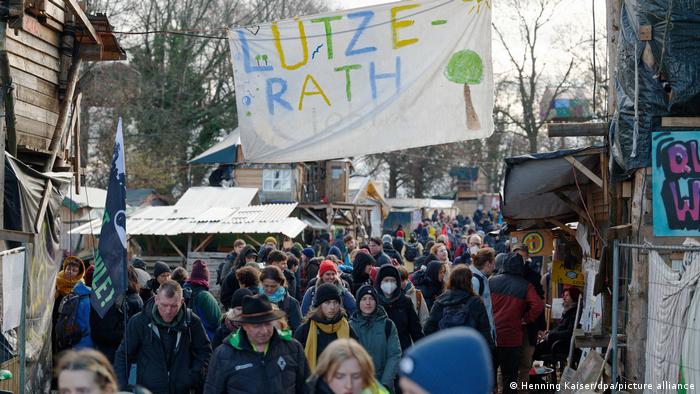 Viele Menschen ziehen durchs Klimacamp von Lützerath, im Hintergrund einige Baumhäuser und Plakate am 8.1.2023