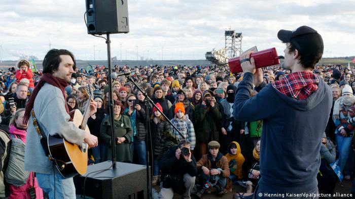Zwei Mitglieder der Band AnnenMayKantereit auf der Bühne, davor viele Zuhörer im Hintergrund der Braunkohlebagger von RWE im Tagebau Garzweiler und die Kohlekraftwerke 
