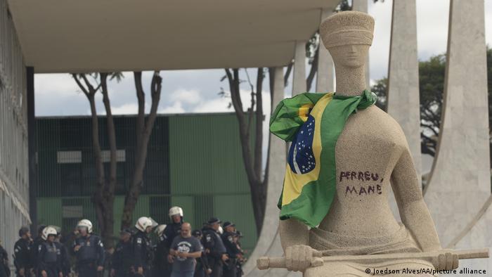 Die Skulptur A Justica - Protestler haben ihr eine brasilianische Flagge umgehängt und einen Spruch auf den Oberkörper geschrieben. Dahinter Sicherheitskräfte.