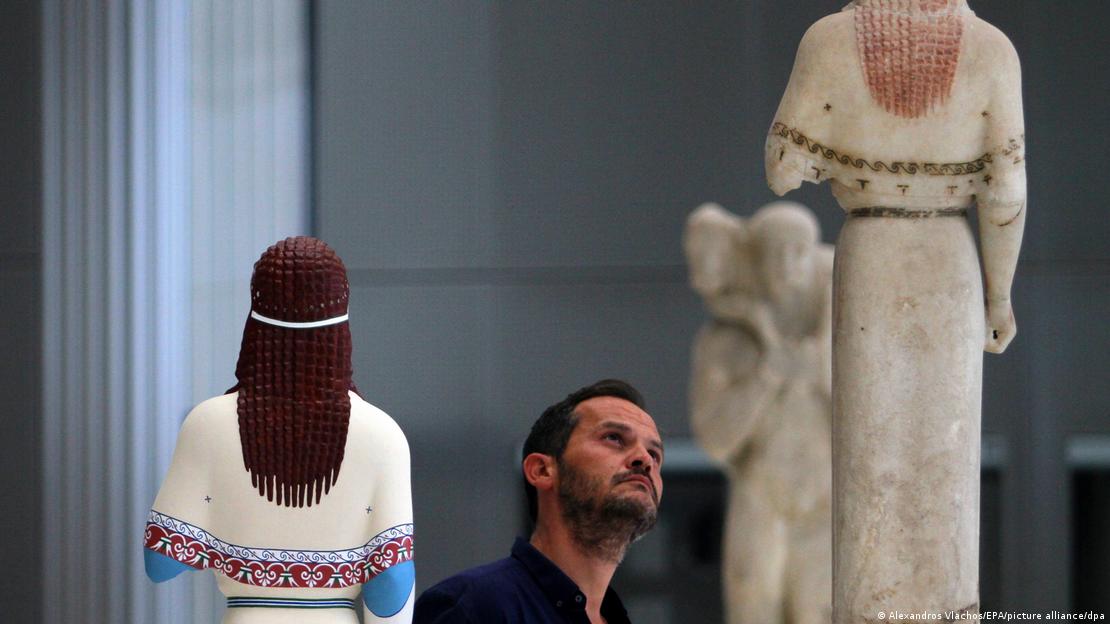 A man looks at a statue of 'The Peplos Kore' next to a colored replica of the same statue.
