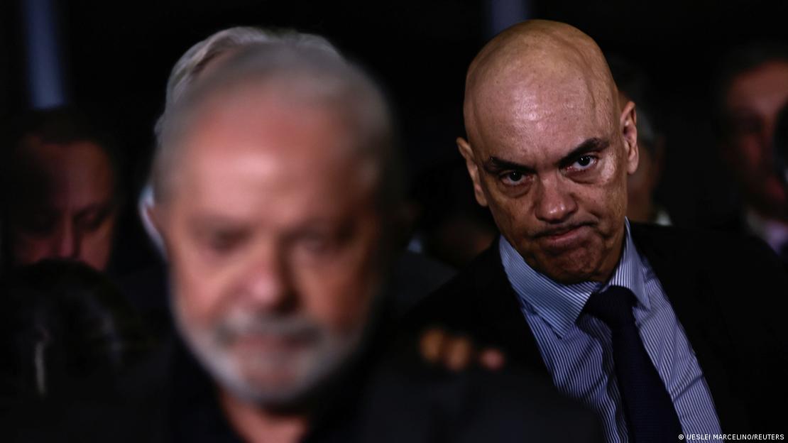 Moraes em segundo plano, com rosto tenso. Em primeiro plano, desfocado, Lula.