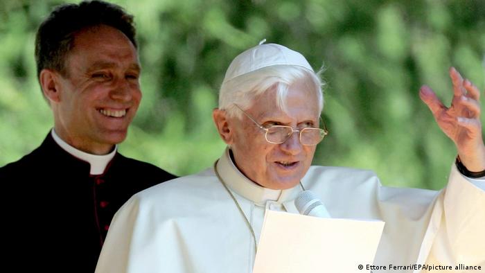 Georg Gänswein steht lachend hinter Papst Benedikt XVI. bei einer Rede