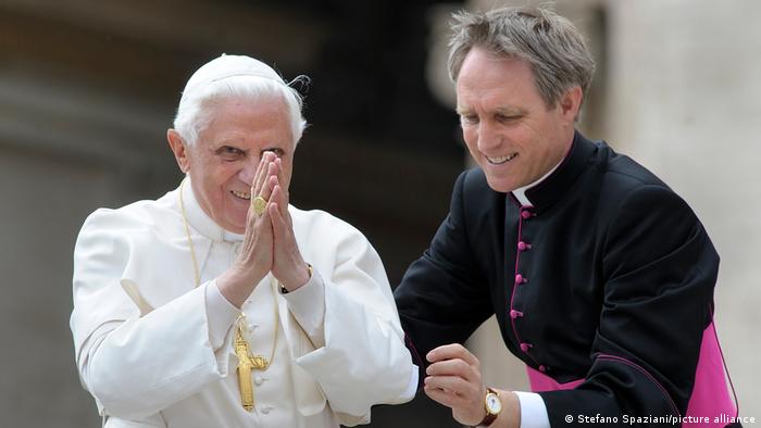 Papst Benedikt faltet die Hände zusammen und grüßt ein Publikum; neben ihm Georg Gänswein 