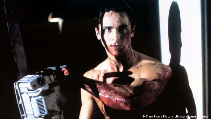 Dieses Filmstill aus American Psycho zeigt einen blutverschmierten Mann mit nacktem Oberkörper, der einen Schatten an die Wand wirft. Er hält ein Gerät in einer Hand.
