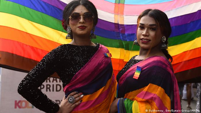 Ein Trans-Paar steht vor einer Regenbogenfahne und hat sich jeweils eine umgelegt