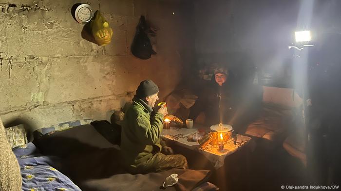 Ein älteres Paar sitzt auf Betten in einem Raum aus rohem Beton an einem Tisch mit und trinkt Tee