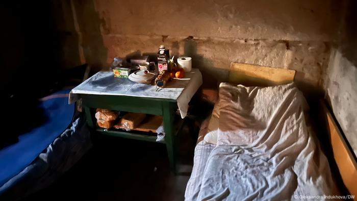Vor einer nackten Steinwand stehen zwei Betten, dazwischen ein kleiner Tisch mit Toastbrot, Tee und weiteren Lebensmitteln