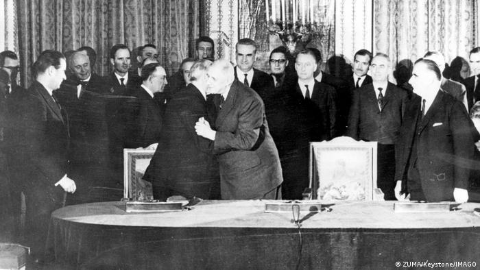 Konrad Adenauer und Charles de Gaulle küssen sich, im Hintergrund stehen viele Männer 