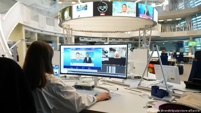 A woman sits in front of a computer screen in a newsroom with several monitors attached to a rack in the middle of the room
