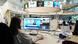 A woman sits in front of a computer screen in a newsroom with several monitors attached to a rack in the middle of the room