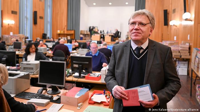 Stephan Bröchler është duke dhënë intervistë, pas tij stafi i tij i ulur përpara kompjuterave duke punuar.