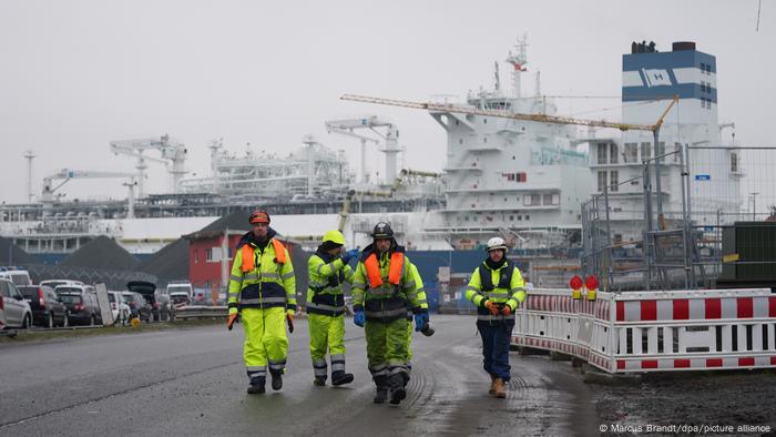 Vier Arbeiter gehen vor dem schwimmenden LNG-Terminal im Industriehafen Brunsbüttel her.