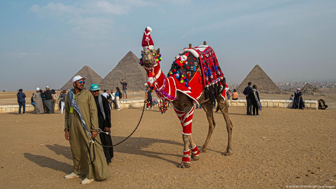 A handler leads his camel, dressed in christmas themed clothing, near the Giza Pyramids.