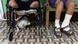 Foto mostra, da cintura para baixo, uma pessoa em uma cadeira de rodas, com as pernas amputadas. Um cachorro está sentado embaixo da cadeira de rodas. Do lado, há um homem com pés enfaixados. Foto mostra, da cintura para baixo, uma pessoa em uma cadeira de rodas, com as pernas amputadas. Um cachorro está sentado embaixo da cadeira de rodas. Do lado, há um homem com pés enfaixados.