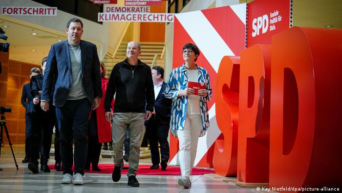 Bundeskanzler Olaf Scholz (SPD), Lars Klingbeil (l), Bundesvorsitzender der SPD, und Saskia Esken (r), Bundesvorsitzende der SPD, gehen durch das Atrium des Willy-Brandt-Hauses in Berlin. Am Rand ist eine große Installation aus den Buchstaben SPD in rot zu sehen.