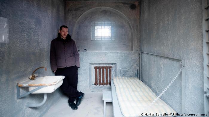 Eine Person steht in einer nachgebauten Einzelzelle aus Beton mit einem Klappbett, einem Waschbecken, einer kleinen Heizung und einem kleinen vergitterten Fenster