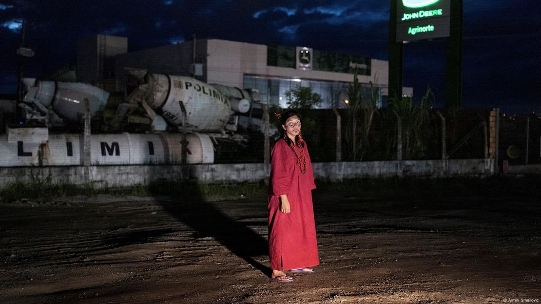 Woman in a red dress stands in a muddy plot, behind her a fenced-in building